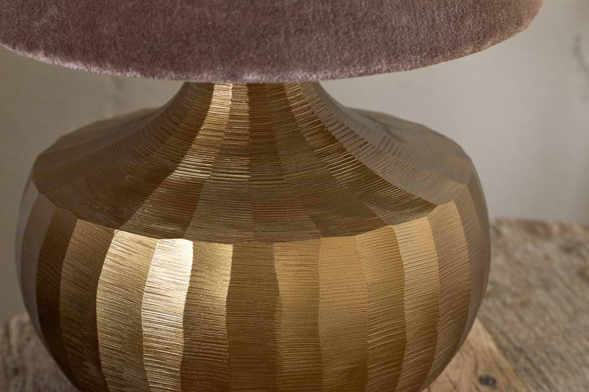 Close-up of the Sakiti Cast Aluminium Table Lamp by nkuku in antique brass, featuring a textured base with vertical ridges and topped with a soft, plush mauve lampshade on a rustic wooden surface.