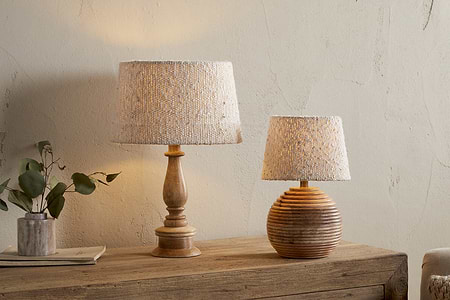 Two Sakiti Bouclé Lampshade – Off White by nkuku lamps add ambient light atop a rustic dresser. One lamp has a tall carved wooden base, the other a round ribbed base. A vase with green leaves and a tray sit against the cream wall.