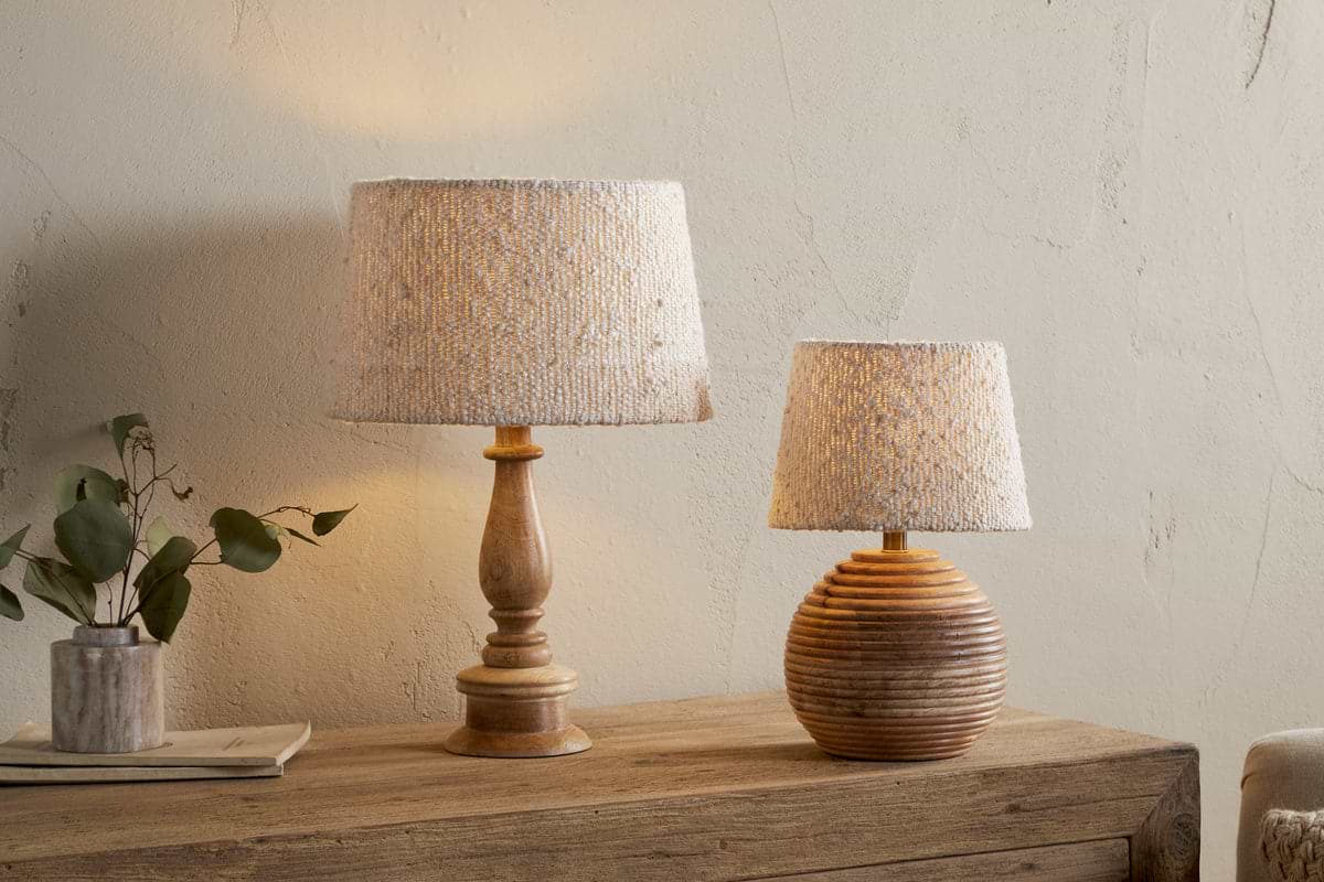 Two Sakiti Bouclé Lampshade – Off White by nkuku lamps add ambient light atop a rustic dresser. One lamp has a tall carved wooden base, the other a round ribbed base. A vase with green leaves and a tray sit against the cream wall.