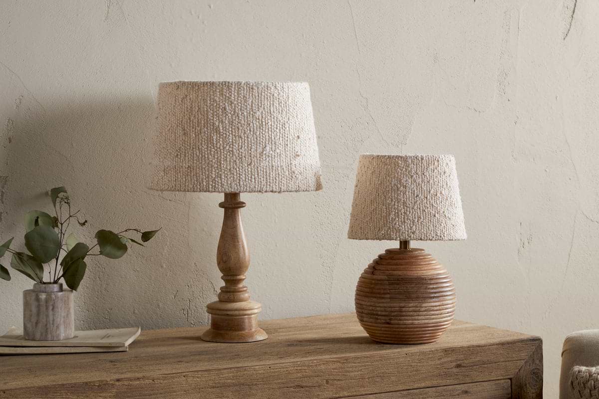 Two nkuku Sakiti Bouclé Lampshades in off white provide soft lighting on a rustic wooden table, each with a unique base. A small vase with greenery rests nearby, set against a textured off-white wall.