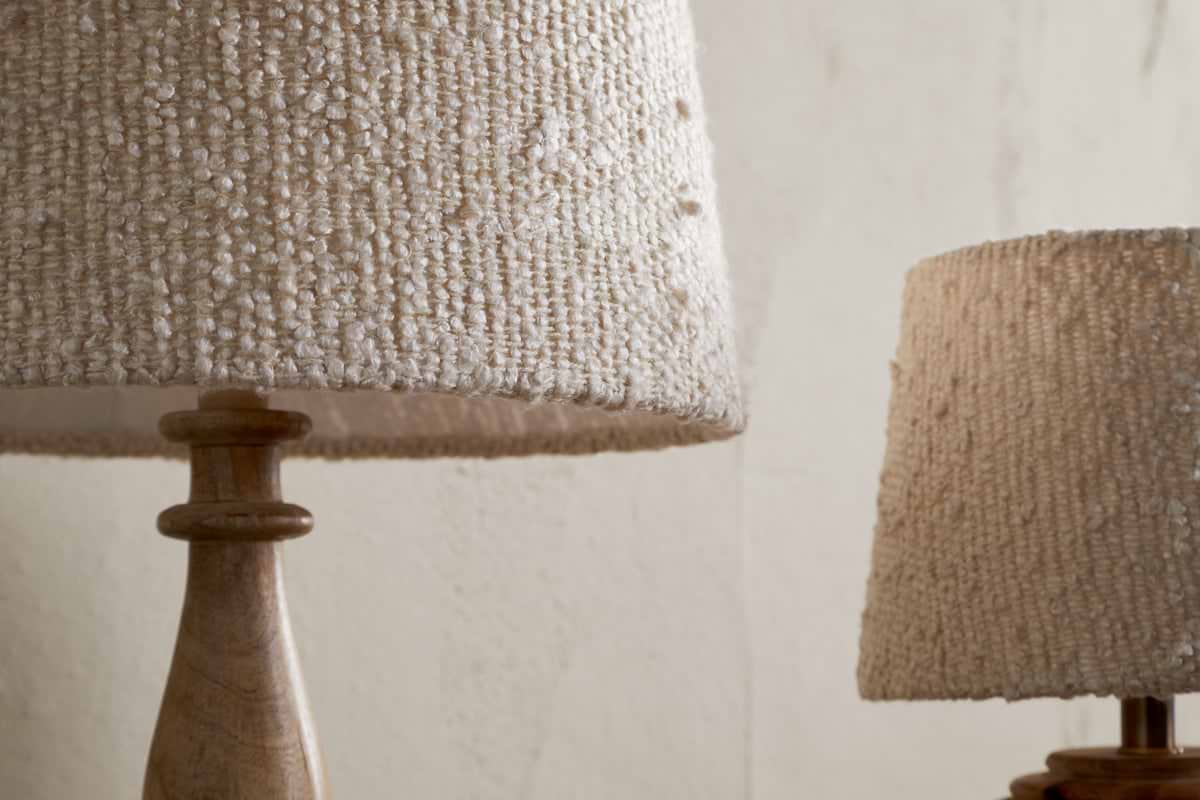Close-up of two nkuku Sakiti Bouclé Lamps with off-white, textured woven lampshades and wooden bases. One lamp stands prominently in the foreground, while the other is partially visible against a neutral wall.