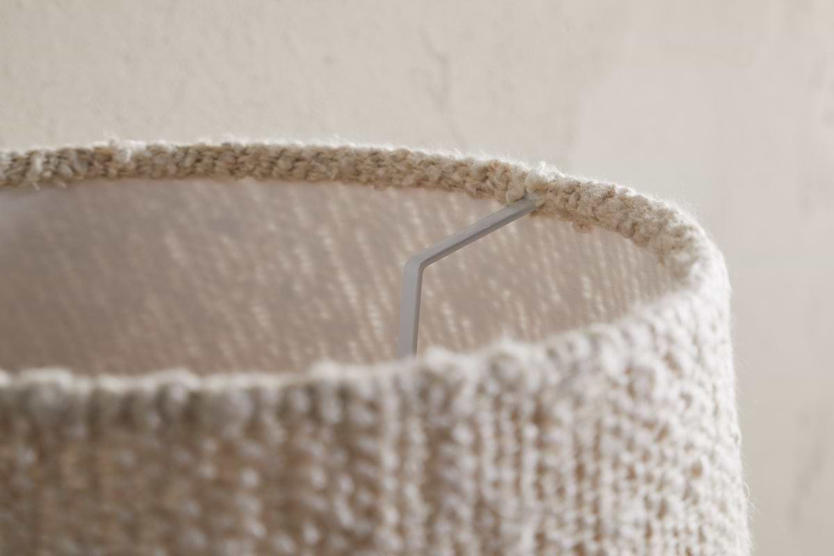 Close-up of the top edge of the Sakiti Bouclé Lampshade in off white by nkuku, featuring textured woven fabric. A metal support arm is visible inside, with soft ambient lighting casting gentle shadows on its surface and the wall.