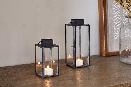 Two nkuku Sia Mini Table Lanterns in black, each holding a lit tealight, are arranged on a wooden surface beside a framed artwork and a glass jar of dried flowers.