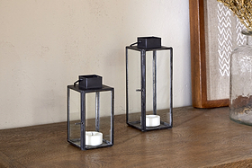 Two nkuku Sia Mini Table Lanterns in black glass and metal—one short, one tall—rest on a wooden surface, each holding a white tealight. Decorative items appear partially in the background.
