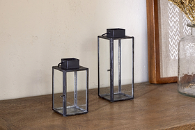 Two Sia Mini Table Lanterns by nkuku, featuring black metal frames, rest on a wooden surface near a glass vase and framed artwork—perfect for holding tealights and enhancing your decor.