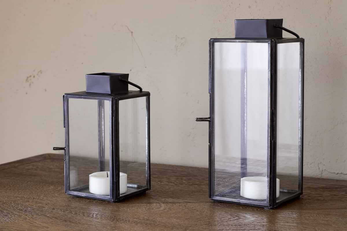 Two nkuku Sia Mini Table Lanterns in black, featuring antique-style frames and glass panes, sit on a wooden surface with white tealights inside. One lantern is taller than the other, set against a beige wall.