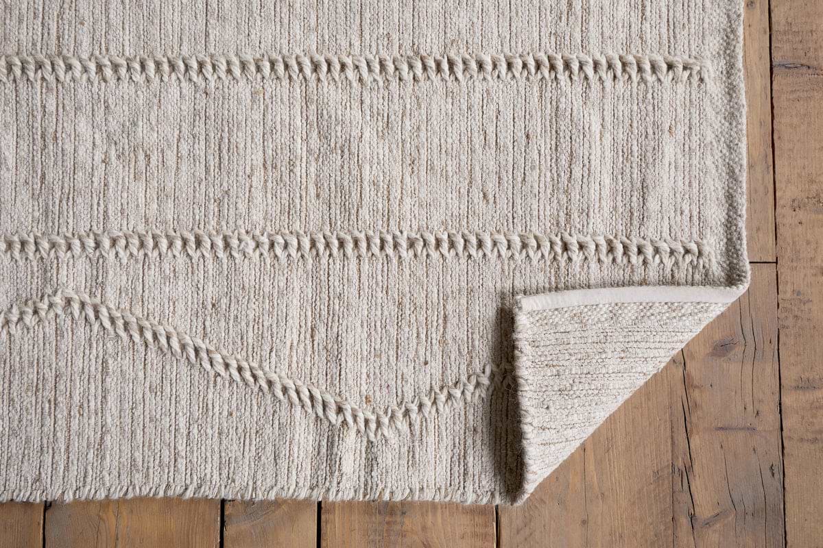 The Shanti Wool & Cotton Rug by nkuku in natural is shown on a wooden floor with one corner folded to display its textured geometric pattern, detailed edging, and a glimpse of the underside.