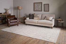 A cozy living room with a beige sofa, brown leather armchair, neutral pillows, wooden side tables, decorative wall art, wooden floors, and the nkuku Shanti Wool & Cotton Rug in Natural.