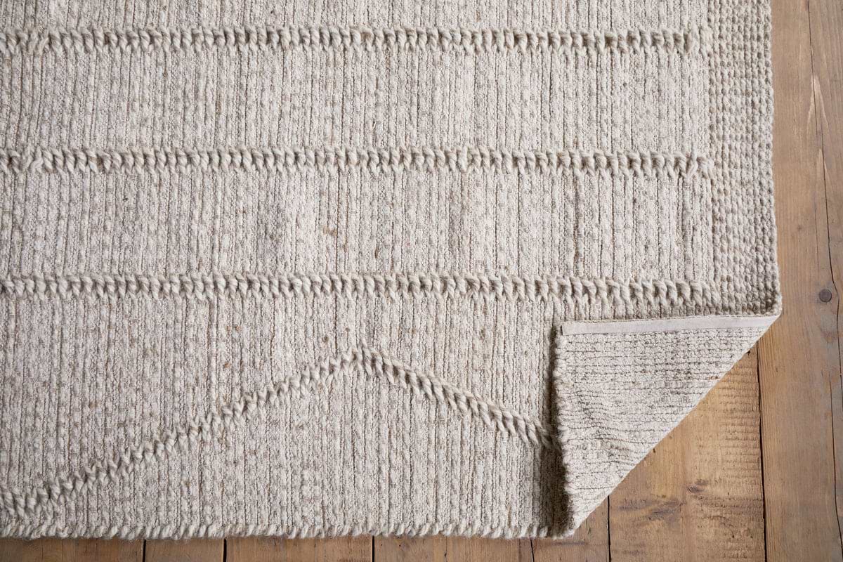 The Shanti Wool & Cotton Rug - Natural by nkuku, featuring subtle geometric patterns, is spread on a wooden floor with its bottom right corner folded to reveal the stylish underside.