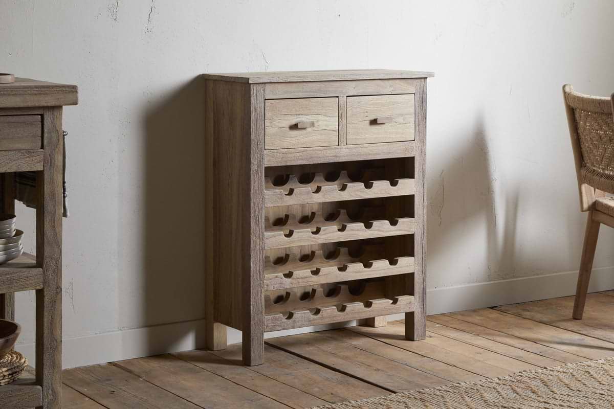 The Shanthi Reclaimed Wood Wine Rack - Natural by nkuku stands against a white wall on a wooden floor, featuring two small drawers and horizontal slots for wine storage. A woven chair and other furniture are partially visible nearby.