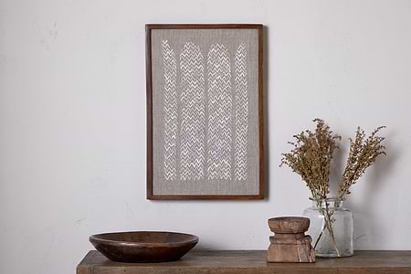The Samaya Hand Stitched Wall Art - Natural by nkuku, featuring a white zigzag on beige, hangs above a wooden surface with a sustainable mango wood bowl, two stacked wooden pieces, and a glass jar of dried flowers.