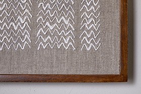 A close-up of the Samaya Hand Stitched Wall Art - Natural by nkuku, showing hand-stitched white zigzags on beige fabric in a sustainable mango wood frame, with the frame’s lower right corner and wall edge visible.