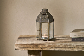 The nkuku Sabra Lantern - Aged Antique, with a glowing tealight inside, rests on a rustic wooden table beside two stacked books. The plain beige wall behind complements the lantern’s cozy, simple atmosphere.