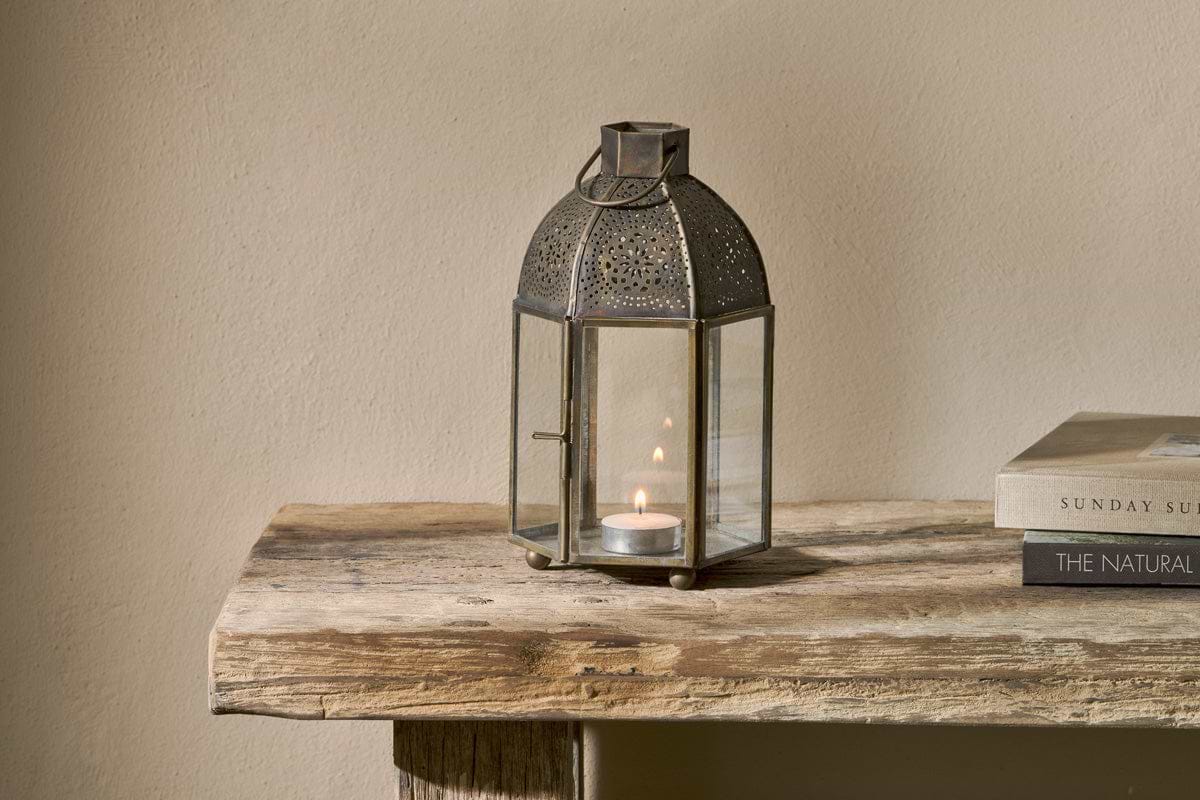 The nkuku Sabra Lantern - Aged Antique, with a glowing tealight inside, rests on a rustic wooden table beside two stacked books. The plain beige wall behind complements the lantern’s cozy, simple atmosphere.