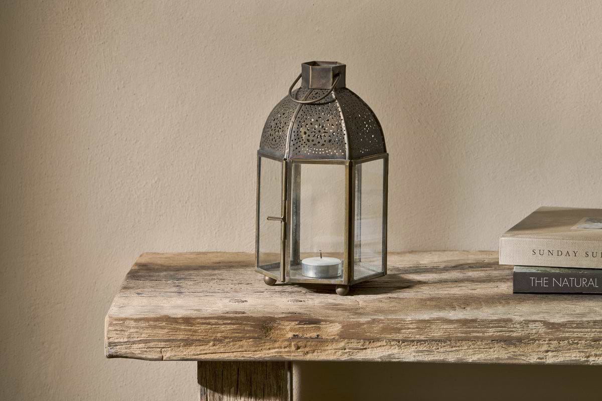 The nkuku Sabra Lantern - Aged Antique with a small tealight holder rests on a rustic wooden table next to two stacked books, while the plain beige wall behind enhances the cozy, minimalist atmosphere.