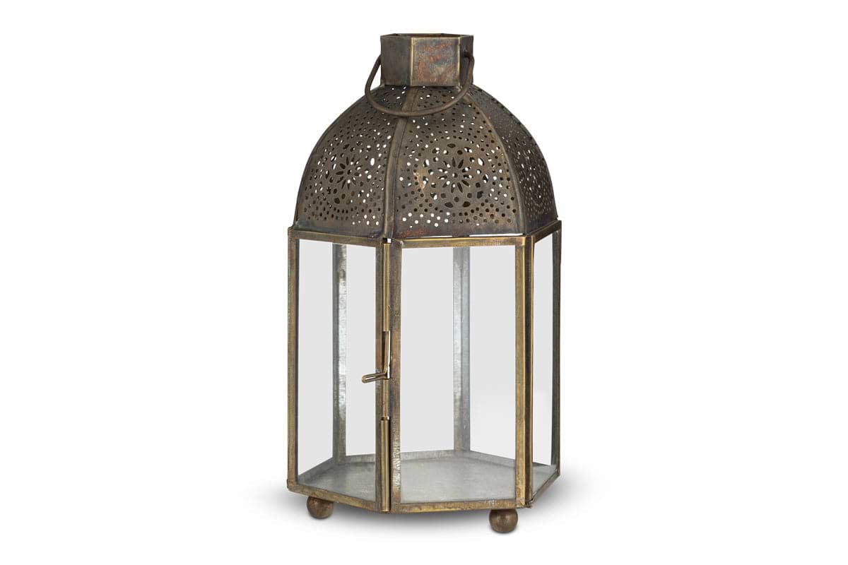 The Sabra Lantern - Aged Antique by nkuku is a hexagonal metal tealight holder with glass panels, a hinged door, latch, domed top with cut-out patterns, round handle, ball feet, and an antique finish for vintage charm.