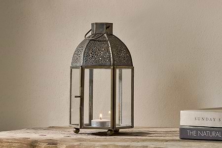 The nkuku Sabra Lantern - Aged Antique features glass panels and an antique finish. A candle glows inside, creating a cozy vibe on a rustic wooden surface, beside two closed books—one titled THE NATURAL.
