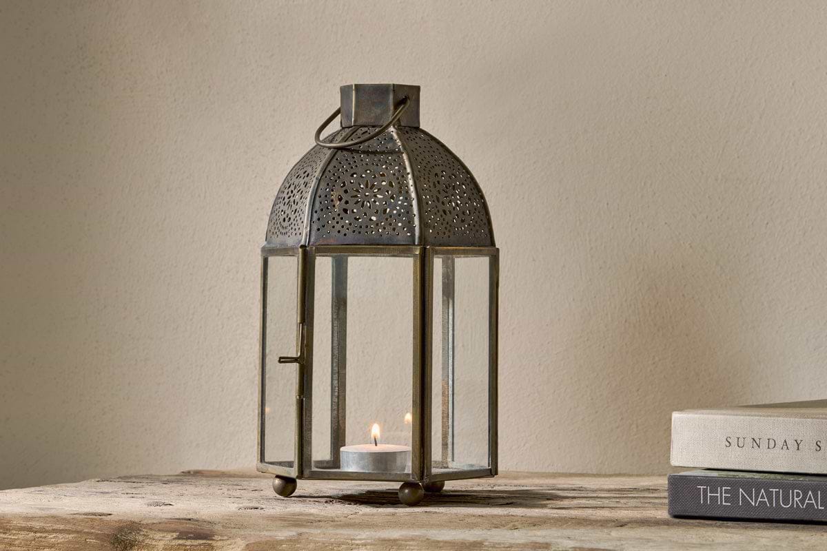 The nkuku Sabra Lantern - Aged Antique features glass panels and an antique finish. A candle glows inside, creating a cozy vibe on a rustic wooden surface, beside two closed books—one titled THE NATURAL.