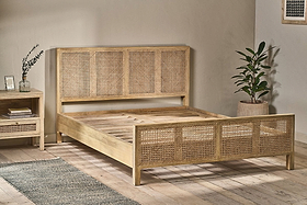 A minimalist bedroom showcases the nkuku Saharsa Cane & Mango Wood Bed, featuring a handwoven cane headboard and footboard. A matching nightstand, potted plant, glass vase with leaves, framed art, light wood floors, and a gray rug complete the space.