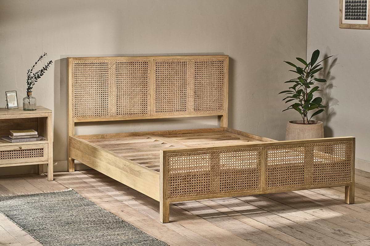 A minimalist bedroom showcases the nkuku Saharsa Cane & Mango Wood Bed, featuring a handwoven cane headboard and footboard. A matching nightstand, potted plant, glass vase with leaves, framed art, light wood floors, and a gray rug complete the space.