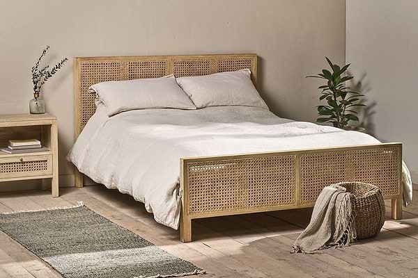 A cozy bedroom features the Saharsa Cane & Mango Wood Bed by nkuku, with a handwoven cane headboard and footboard dressed in white bedding. A matching nightstand holds a vase and books, while a green plant and basket sit on the gray rug.
