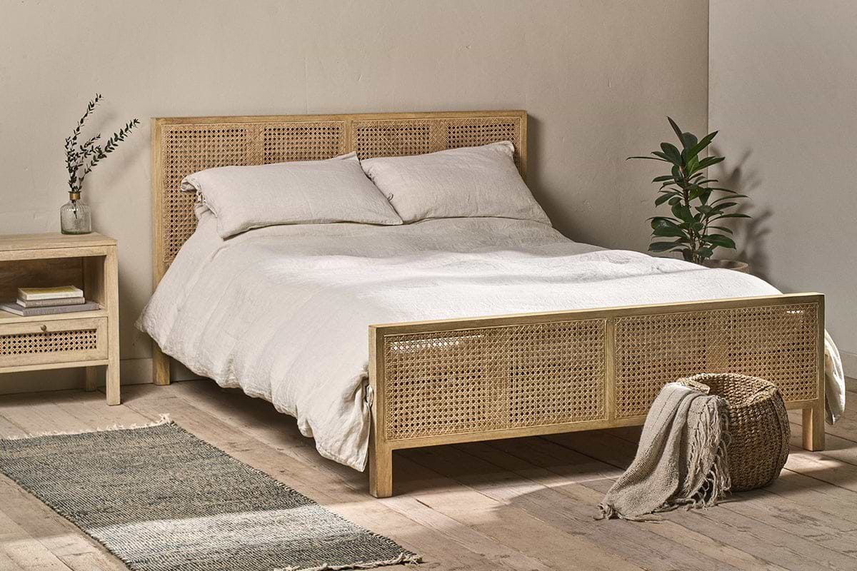 A cozy bedroom features the Saharsa Cane & Mango Wood Bed by nkuku, with a handwoven cane headboard and footboard dressed in white bedding. A matching nightstand holds a vase and books, while a green plant and basket sit on the gray rug.