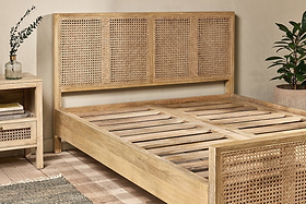 The nkuku Saharsa Cane & Mango Wood Bed, featuring a handwoven cane headboard and sustainable mango wood frame, is paired with a matching nightstand. A plant decorates the background.