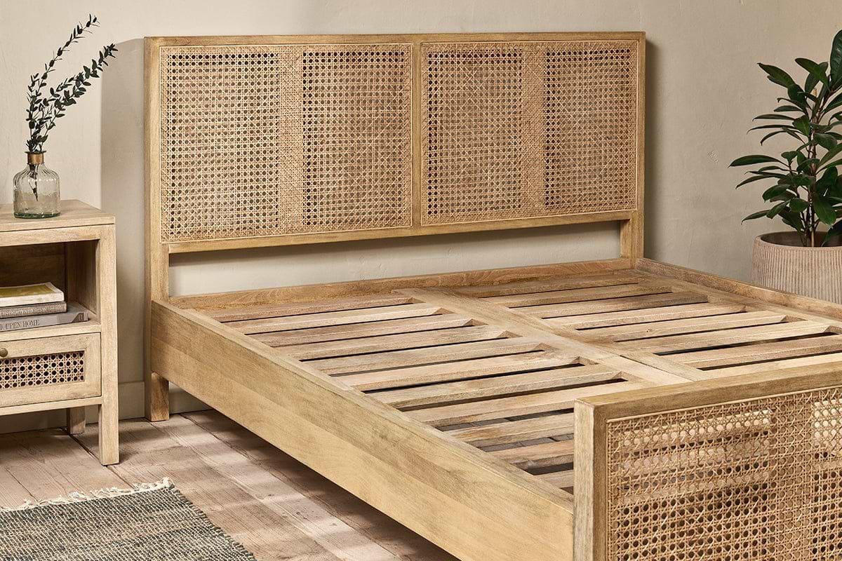 The nkuku Saharsa Cane & Mango Wood Bed, featuring a handwoven cane headboard and sustainable mango wood frame, is paired with a matching nightstand. A plant decorates the background.