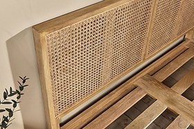 The nkuku Saharsa Cane & Mango Wood Bed features a close-up of its light natural-finish mango wood frame and handwoven cane headboard. The slatted base is visible against a beige background with a sprig of greenery in the corner.