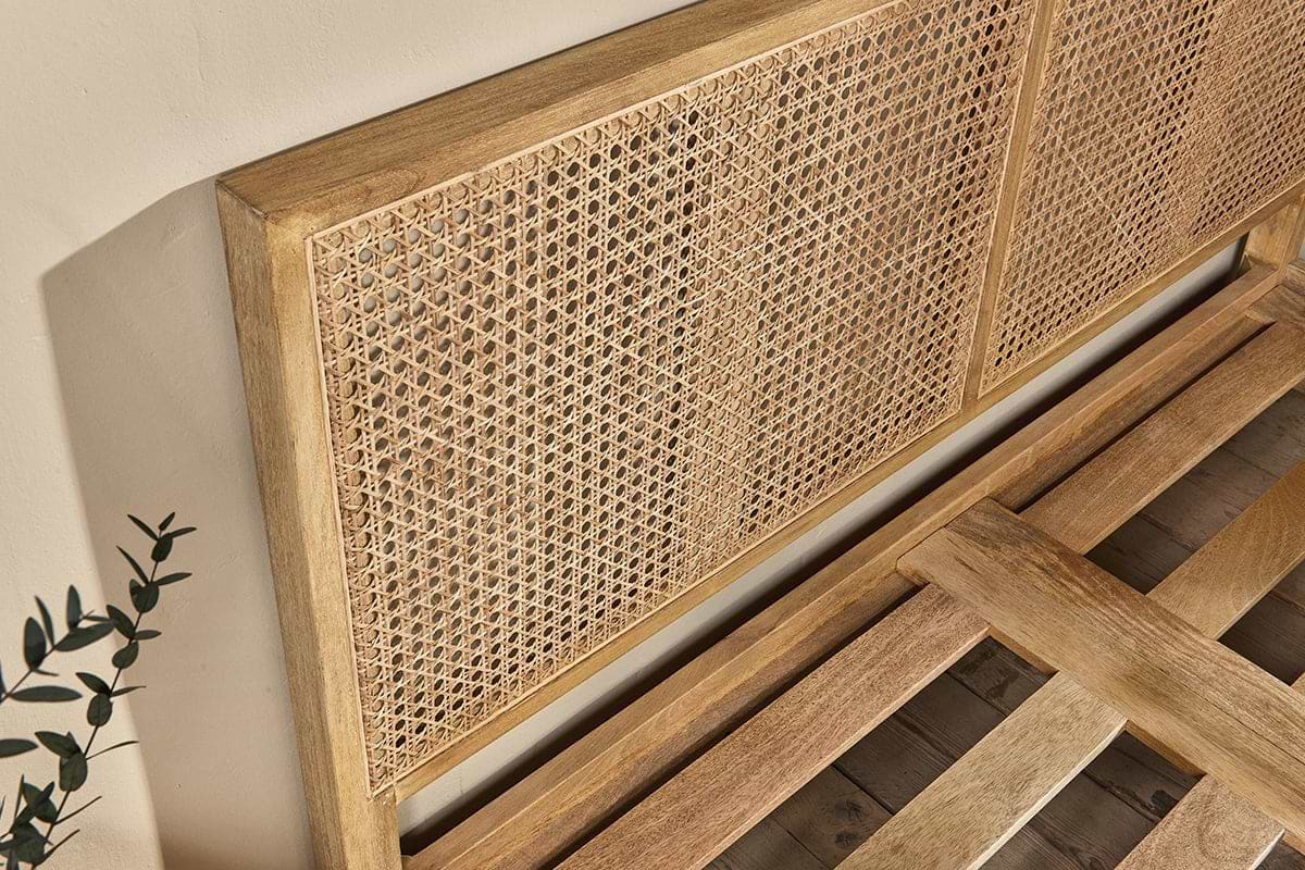 The nkuku Saharsa Cane & Mango Wood Bed features a close-up of its light natural-finish mango wood frame and handwoven cane headboard. The slatted base is visible against a beige background with a sprig of greenery in the corner.