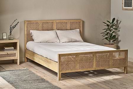 The nkuku Saharsa Cane & Mango Wood Bed, featuring handwoven cane details, is styled with white linens and pillows, and placed between a nightstand and a potted plant.