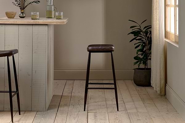 A minimalist room features a wooden bar counter, the nkuku Samaira Leather Counter Stool in dark brown, an ergonomic chair, light wood floors, a potted plant by a sunlit window with sheer curtains, and glass decor in calm neutrals.