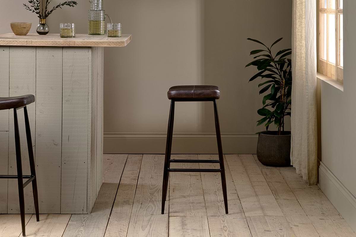 A minimalist room features a wooden bar counter, the nkuku Samaira Leather Counter Stool in dark brown, an ergonomic chair, light wood floors, a potted plant by a sunlit window with sheer curtains, and glass decor in calm neutrals.