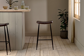 A modern bar area with a light wood counter features two nkuku Samaira Leather Counter Stools in dark brown, black metal legs, light wood flooring, a potted plant, soft natural window light, and glasses on the counter.