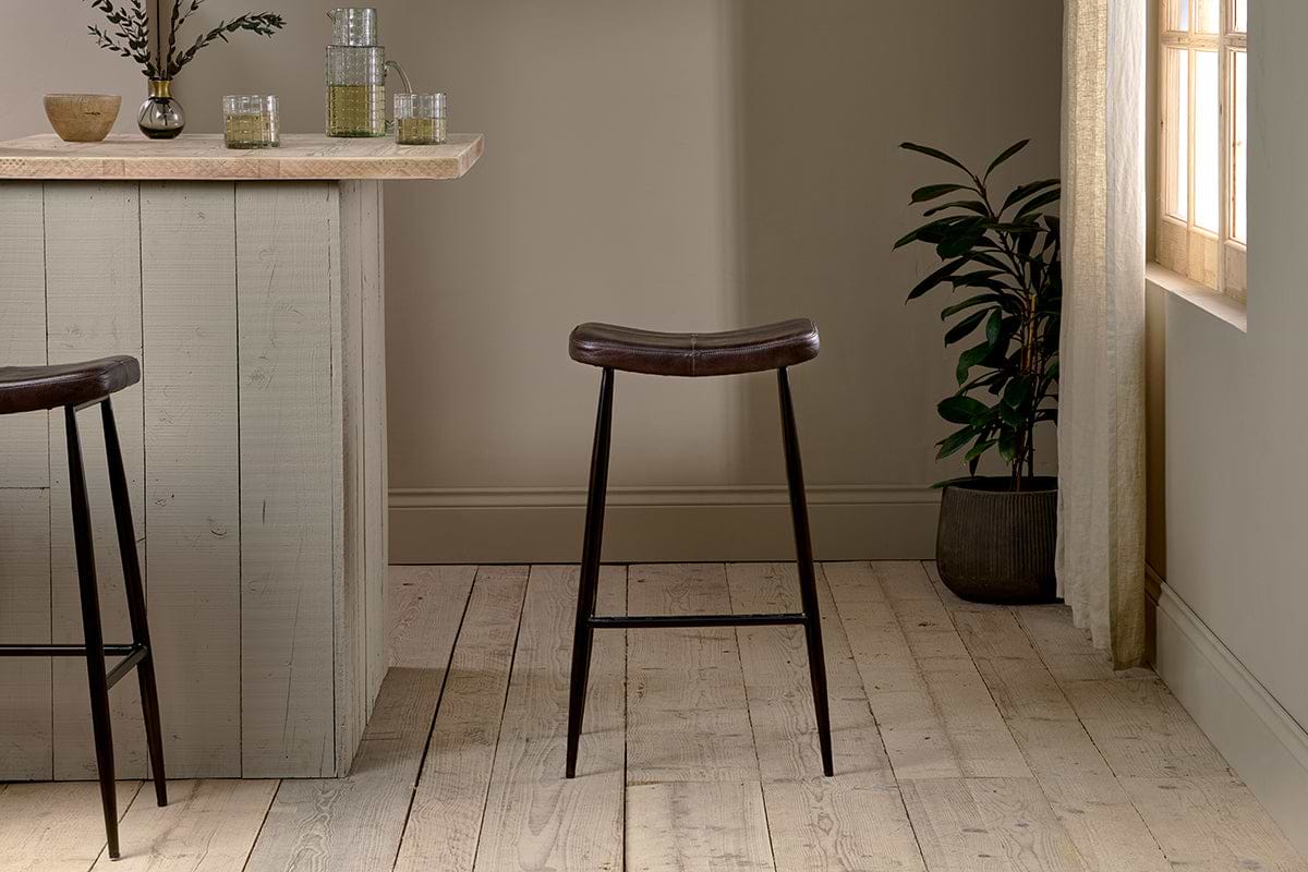 A modern bar area with a light wood counter features two nkuku Samaira Leather Counter Stools in dark brown, black metal legs, light wood flooring, a potted plant, soft natural window light, and glasses on the counter.