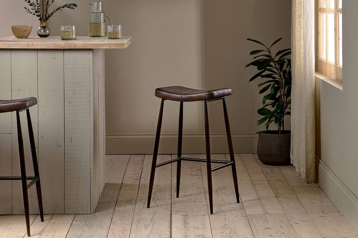 A modern kitchen features a light wood island paired with two nkuku Samaira Leather Counter Stools in dark brown, each with dark metal legs. Glassware and a vase decorate the island, while a potted plant sits by the window with sheer curtains.