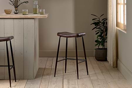 A modern kitchen features a light wood island paired with two nkuku Samaira Leather Counter Stools in dark brown, each with dark metal legs. Glassware and a vase decorate the island, while a potted plant sits by the window with sheer curtains.