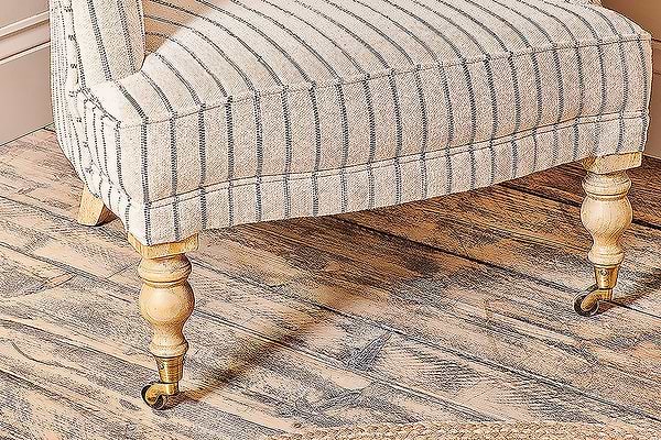 The nkuku Sanja Stripe Armchair, with its curved beige striped fabric and decorative wooden legs featuring brass front casters, sits on a wooden floor with a woven rug edge visible in the corner.