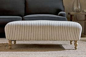 The nkuku Sanja Stripe Upholstered Ottoman in Off White, featuring a natural linen stripe design and four turned wooden legs, sits on a textured rug in front of a dark gray sofa.