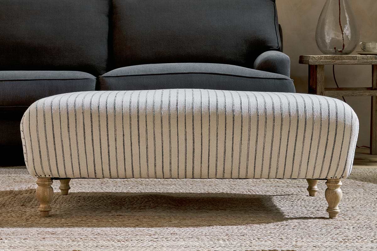 The nkuku Sanja Stripe Upholstered Ottoman in Off White, featuring a natural linen stripe design and four turned wooden legs, sits on a textured rug in front of a dark gray sofa.