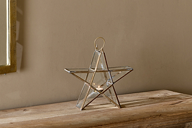 The Sanwi Standing Star Brass by nkuku, crafted from glass and metal with an antique brass finish, holds a small candle on a rustic wooden surface as sunlight from a nearby window casts a soft, warm glow over the minimalist scene.