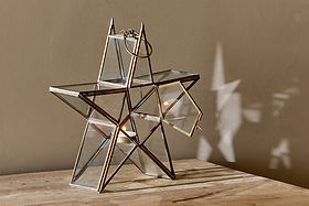 The nkuku Sanwi Standing Star Brass lantern rests on a wooden surface with its side door open, showcasing a glowing tealight. Its glass and brass frame casts a star-shaped shadow on the beige wall behind it.