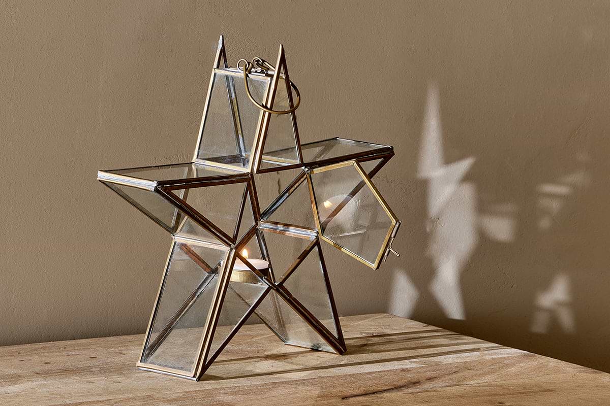 The nkuku Sanwi Standing Star Brass lantern rests on a wooden surface with its side door open, showcasing a glowing tealight. Its glass and brass frame casts a star-shaped shadow on the beige wall behind it.
