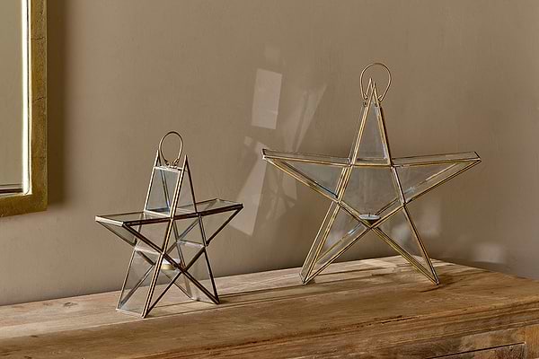 Two Sanwi Standing Star Brass lanterns by nkuku, one small and one large, feature gold-colored metal edges and loop handles. They sit on a wooden surface against a beige wall, casting festive shadows.