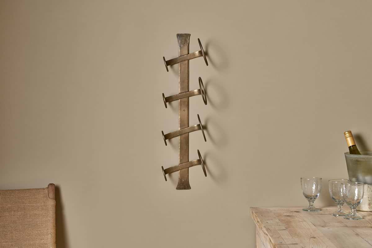 The Shray Iron Wine Rack - Antique Brass by nkuku, featuring four horizontal holders, is mounted on a beige wall above a wooden table with three empty wine glasses, a wine bottle in an ice bucket, and a woven chair partly visible to the left.