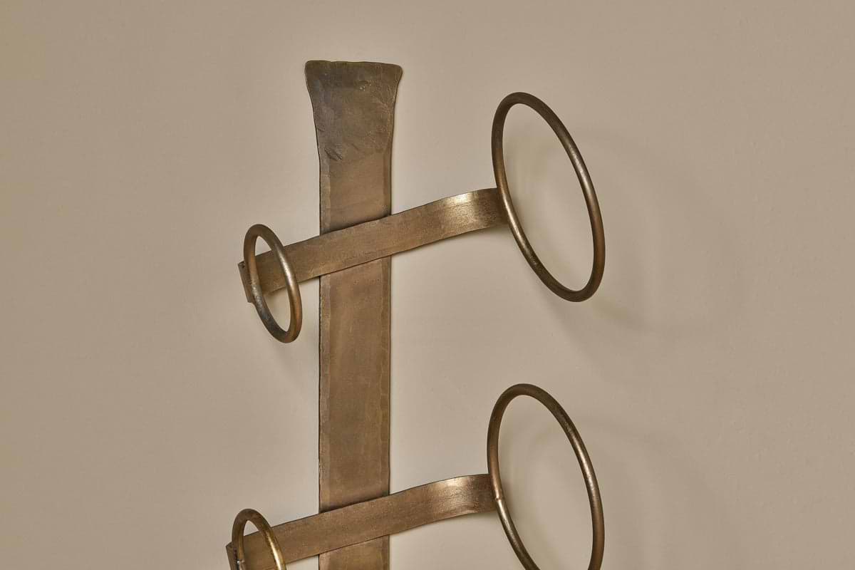 A close-up of the nkuku Shray Iron Wine Rack in antique brass, featuring a vertical strip with three diagonally arranged circular holders, mounted on an off-white wall.