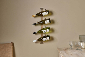 Three wine bottles rest on the nkuku Shray Iron Wine Rack in Antique Brass, mounted on a beige wall above a wooden table with glassware and a pitcher. A woven chair sits nearby, creating a warm, minimalistic setting.