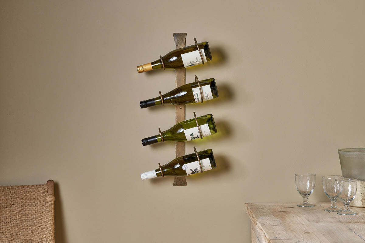Three wine bottles rest on the nkuku Shray Iron Wine Rack in Antique Brass, mounted on a beige wall above a wooden table with glassware and a pitcher. A woven chair sits nearby, creating a warm, minimalistic setting.