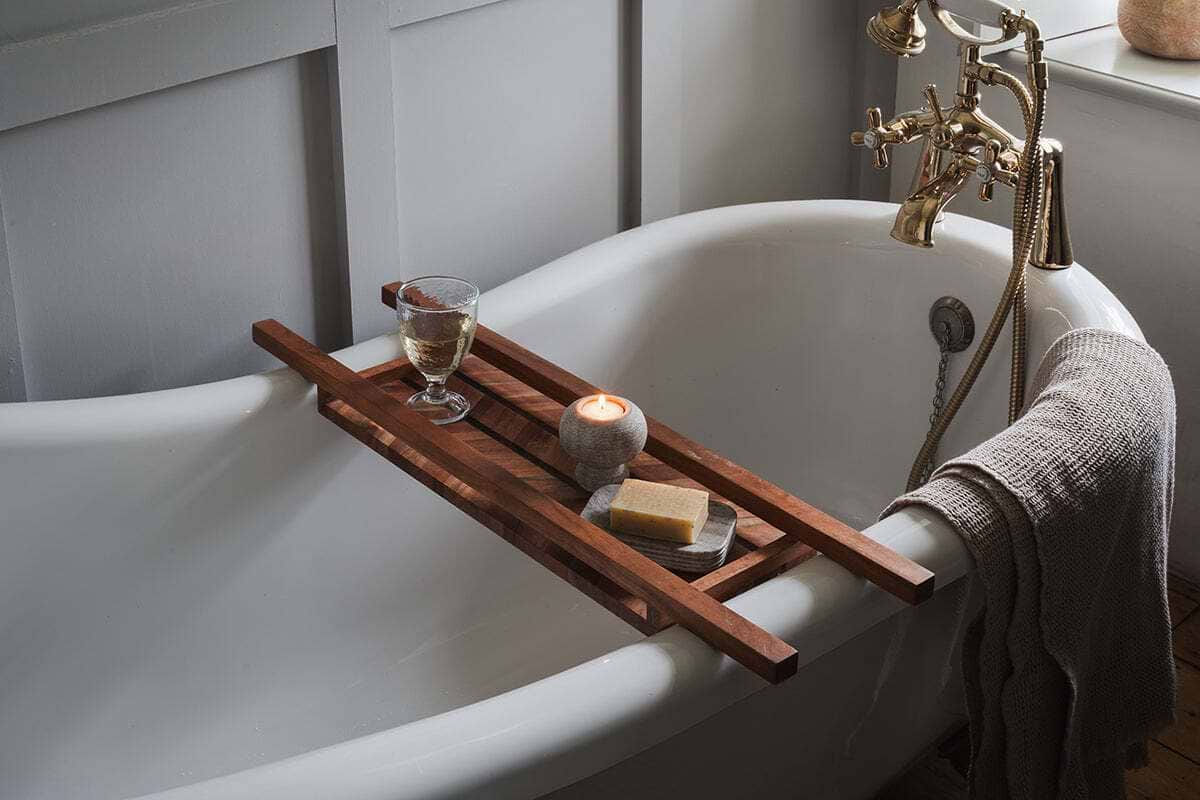 A white clawfoot bathtub features the nkuku Sukha Mango Bath Bridge - Natural holding a lit candle, soap on a dish, and a glass of white wine beside brass fixtures; a beige towel over the side creates a calming, spa-like retreat.