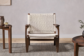 The Surat Woven Lounger - Natural by nkuku features acacia wood, wide armrests, and a hand-woven cotton rope seat and back. Placed on a textured gray rug, it sits between two round tables—one with white wine, the other with a vase of greenery.
